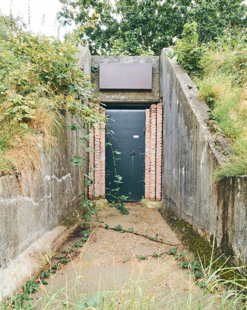 Steenbakke met militaire bunkers bij de Vliegbasis W210, omlijst door groen en natuur, met een blauw metalen deur en bakstenen details, een rustige en historische locatie.
