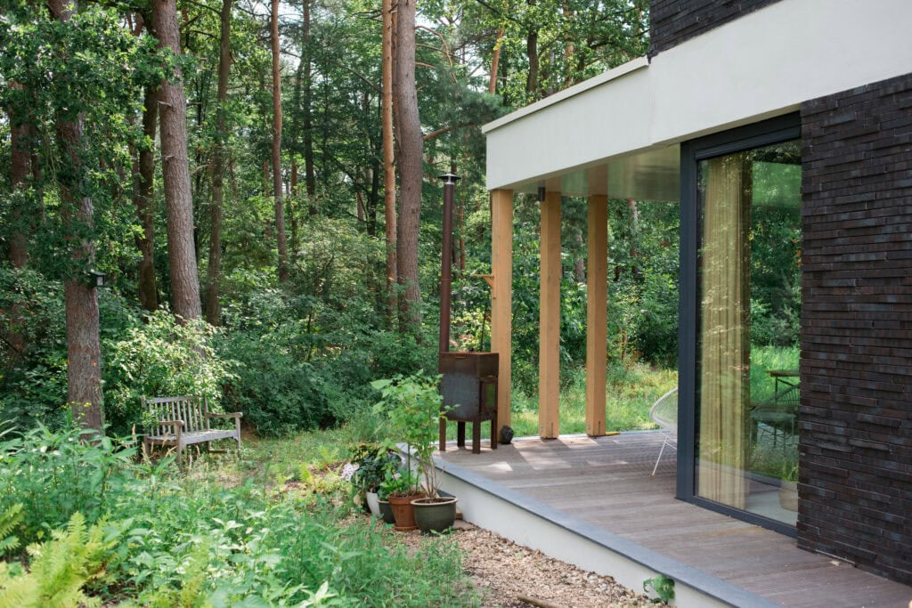 Modern woning met grote glazen gevel en uitzicht op bosrijke omgeving, duurzame bouwmaterialen, houten terras en tuin met planten, perfect voor genieten van natuur en comfort.