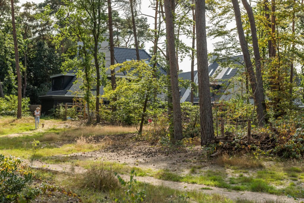 Verzorgde tuin met wandelpad en moderne woningen op de achtergrond in de wijk De Vliegbasis. Uitgestrekte groene bomen en speelse lichtinval bieden een rustige en natuurlijke omgeving.