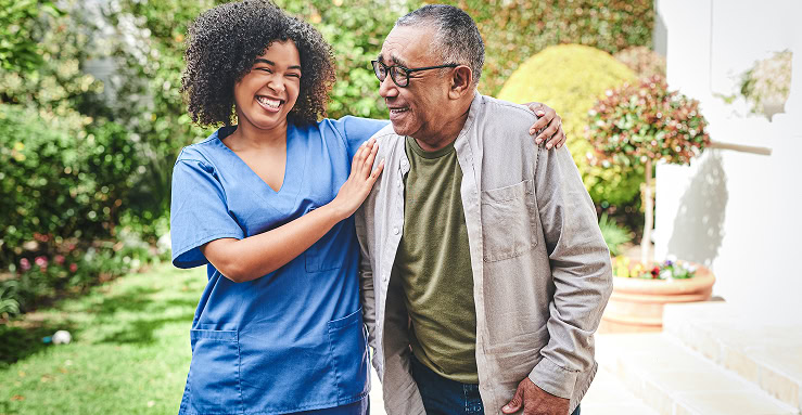 Zorgverlener in blauwe scrubs die zorgbehoevende oudere ondersteunt en vriendelijk glimlacht in een tuin met groene planten en bloemen.