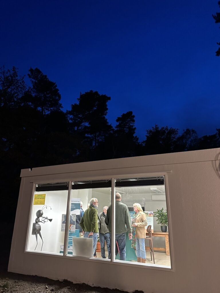 Moderne huisjes op de Vliegbasis met transparante ramen en groepsbijeenkomst, omgeven door bomen onder een heldere blauwe hemel, perfect voor rust en windenergie demonstraties.