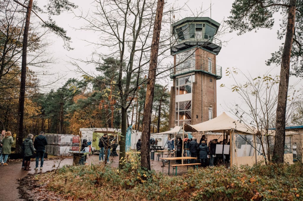 Uitzicht op de toren van de Vliegbasis met bezoekers en marktkraampjes in een bosrijke omgeving tijdens een evenement.