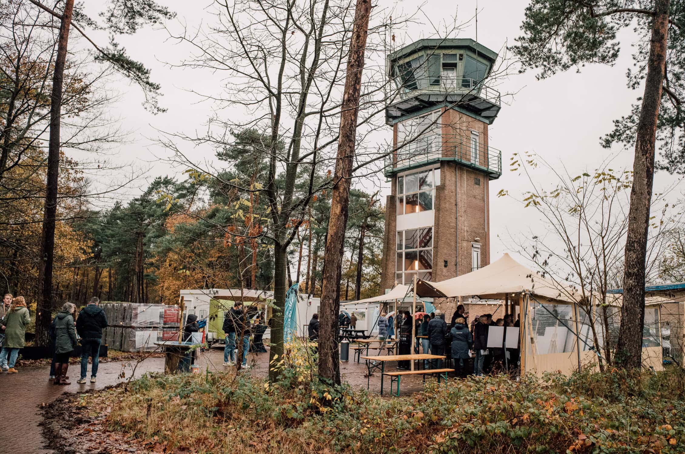 Uitzicht op de toren van de Vliegbasis met bezoekers en marktkraampjes in een bosrijke omgeving tijdens een evenement.