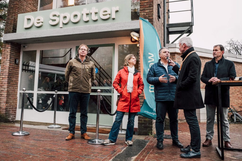 Toegangsdeuren van De Vliegbasis en een bijeenkomst met sprekers voor een evenement op de luchtmachtbasis in Nederland, met een achtergrond van een gebouw met een signaalbord "De Spotter".
