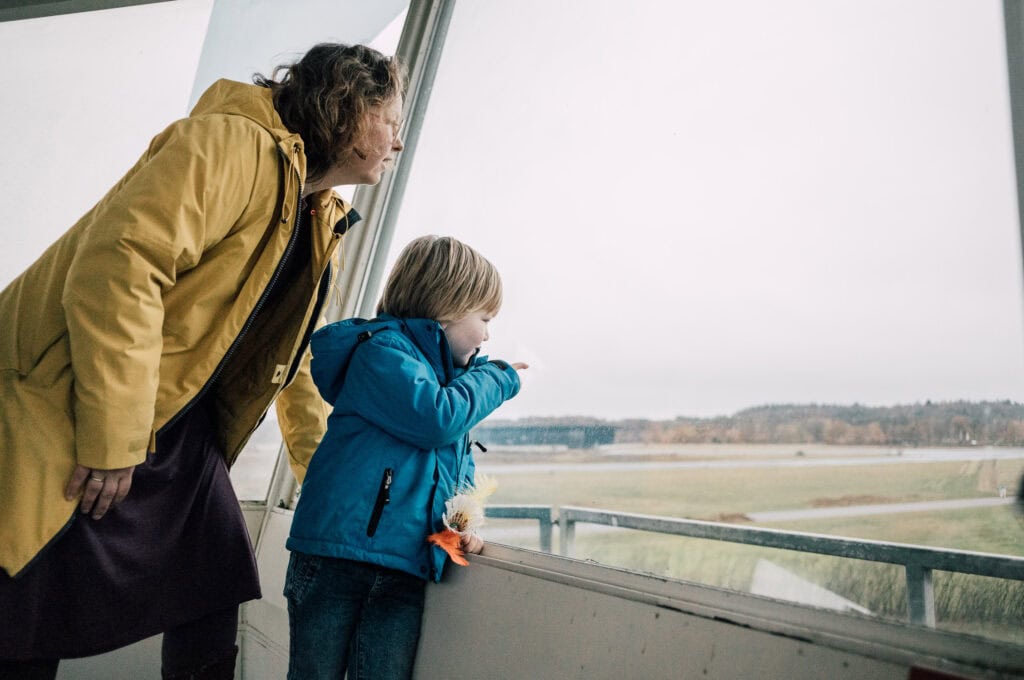 Luchtvaartactiviteit op vliegbasis, familie kijkt uit het uitzicht, kinderen en volwassenen genieten van het vliegverkeer, kind met speelgoedvogel, kind en volwassene in jas bekijken vliegtuigen, omgeving van vliegbasis in Nederland, activiteiten op of rondom de luchtmachtbasis, gezinsuitje op de luchtbasis, verkenning van de luchtvaart in Nederland, observeren van vliegtuigen, familie-uitje op de vliegveldpromenade.