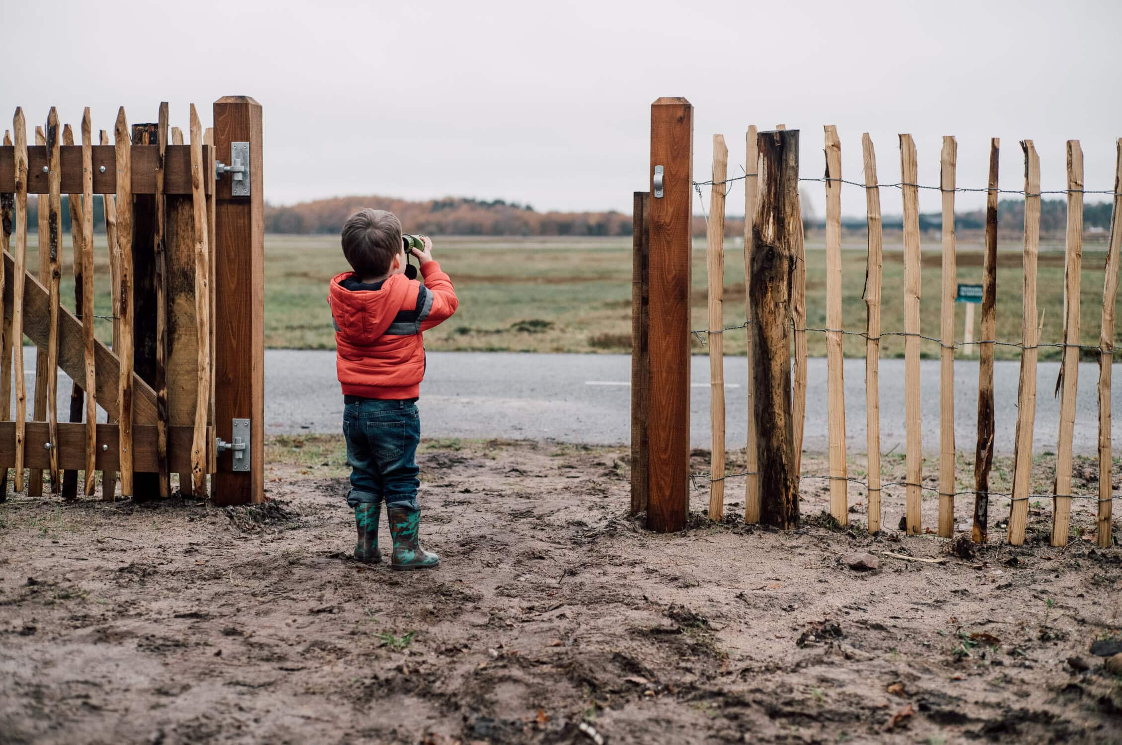 Gezicht van een kind dat door een verrekijker naar de luchtmachtbasis kijkt, met een houten hek op de voorgrond en een uitgestrekte lucht en natuur op de achtergrond.