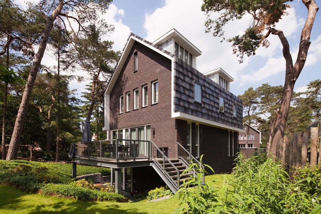 Moderne woning gebouwd op de Vliegbasis, omgeven door groen en bomen, met een unieke architectuur en grote ramen voor natuurlijk licht.