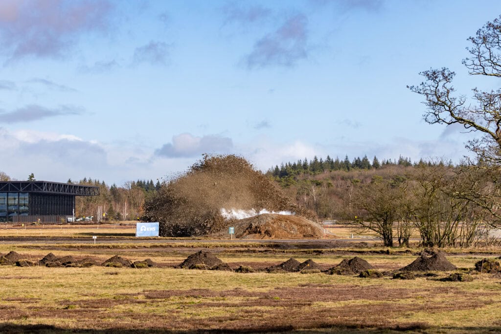 Uitbreiding van de vliegbasis met een grote explosie en stofwolk, omgeven door open velden en enkele bomen, onder een bewolkte blauwe hemel.