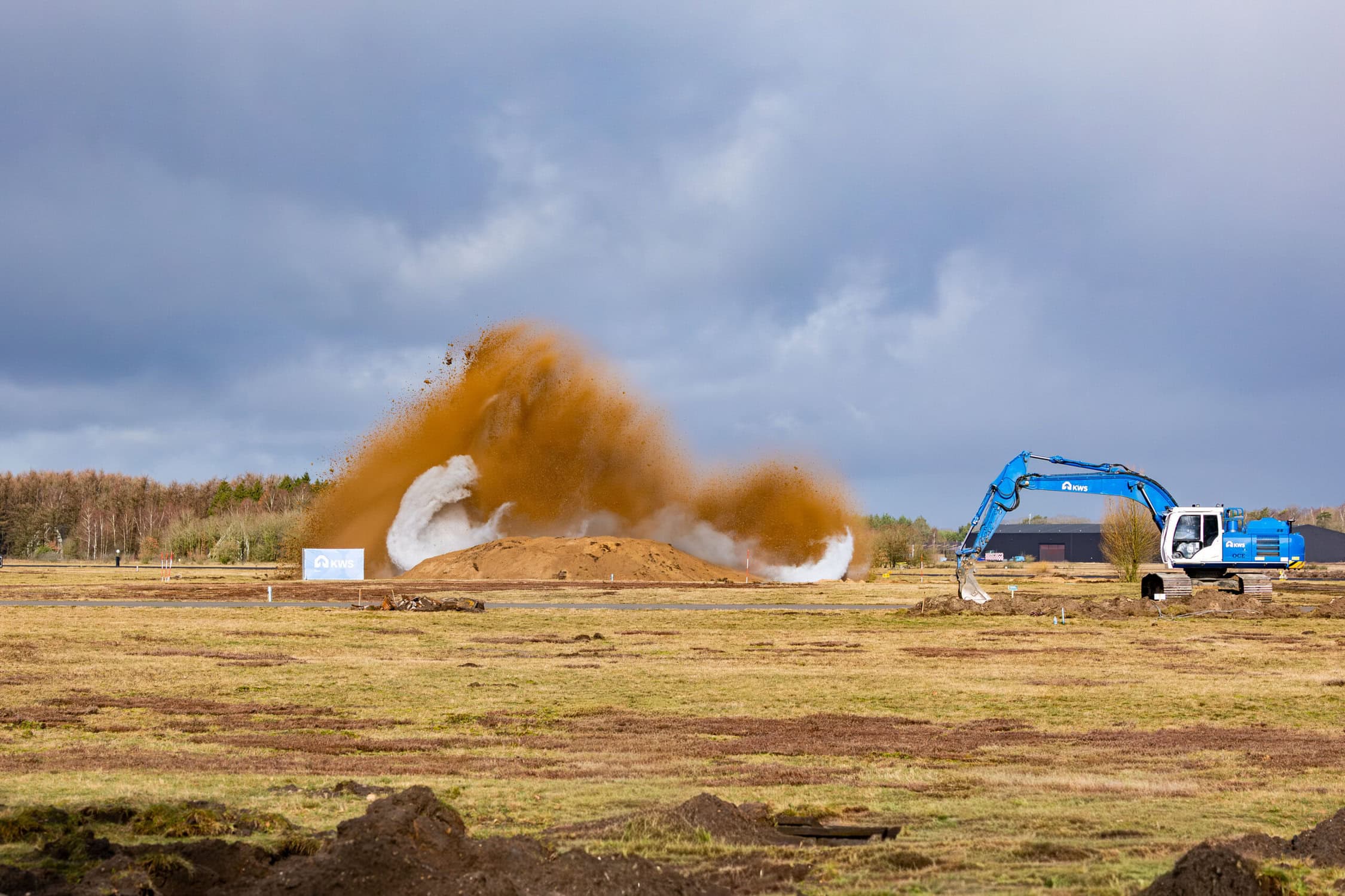 Demonstratie van grondwerk op de Vliegbasis met een graafmachine die aarde verplaatst, onder een bewolkte hemel.