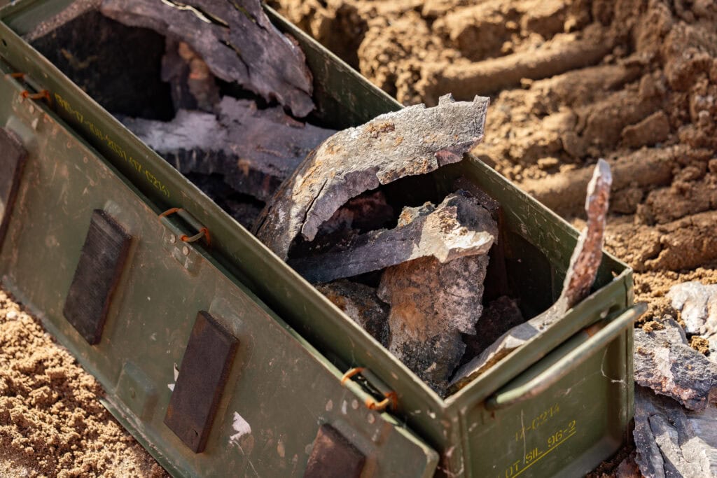Restanten van oorlogsmateriaal in een doos op de grond, met modder en aarde eromheen, op de vliegbasis.