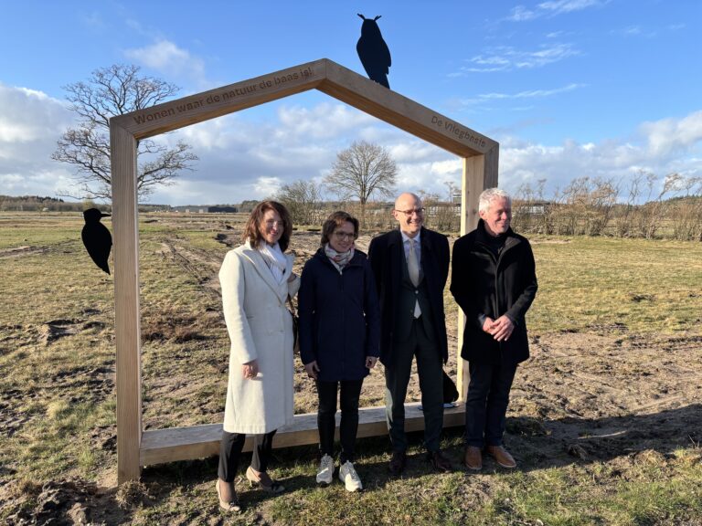 Eurocommissaris bezoekt De Vliegbasis tijdens werkbezoek, omgeven door officials en genodigden, met open veld en blauwe lucht op de achtergrond.