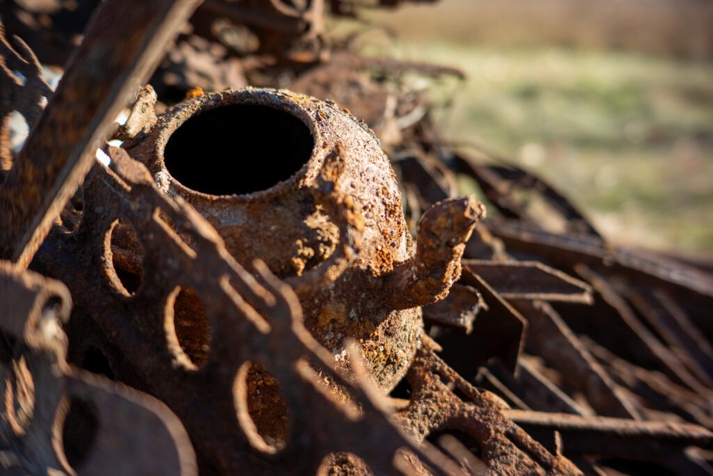 Close-up van een verroeste, oude vliegtuigonderdeel op de vliegbasis, omgeven door roestige metalen onderdelen en afval, met een achtergrond van een open veld.
