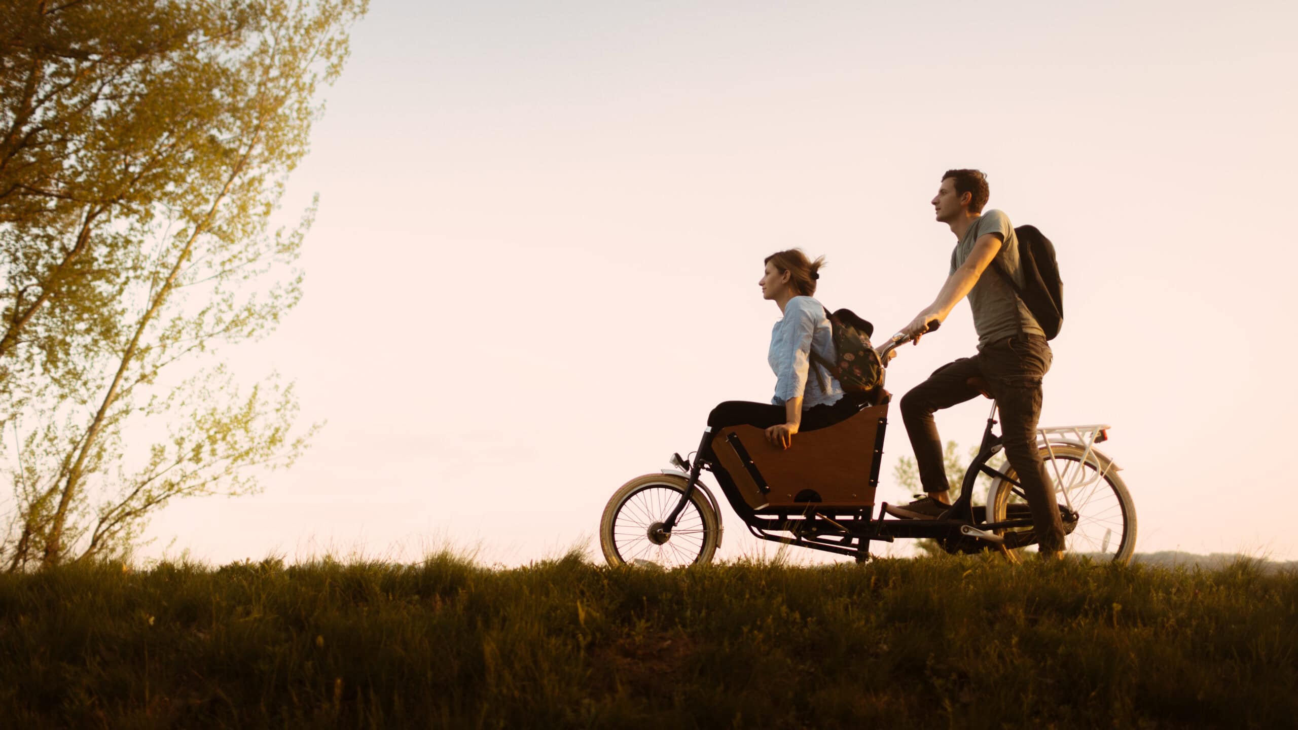 Twee mensen fietsen samen op een tandemfiets in een open landschap bij de Vliegbasis, met een rustige zonsondergang op de achtergrond, onder de titel 'Omgeving'.