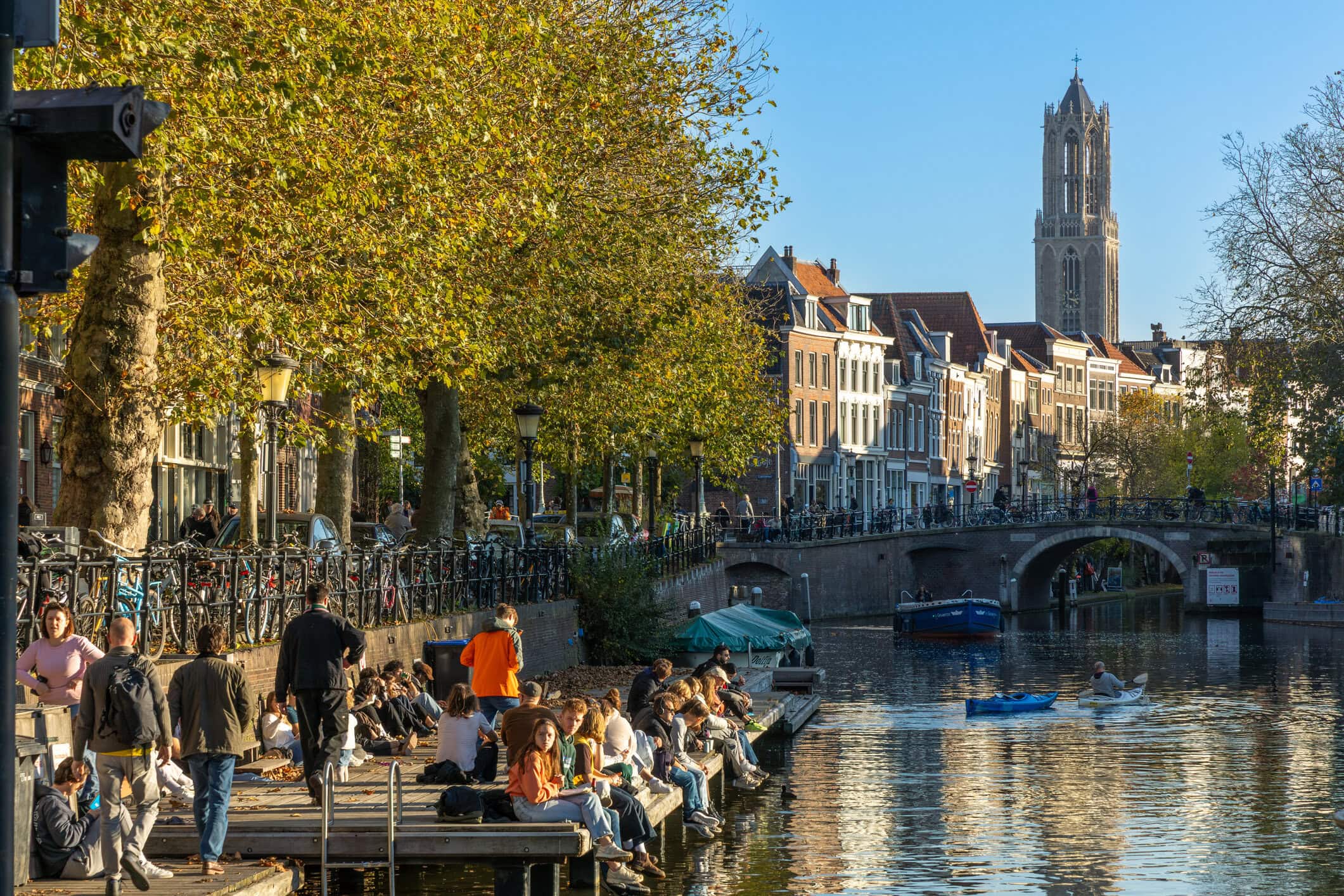 Uitzicht op de omgeving van de Vliegbasis met grachten, bruggen en oude huizen, onder een heldere blauwe hemel, met de toren van een kathedraal op de achtergrond.
